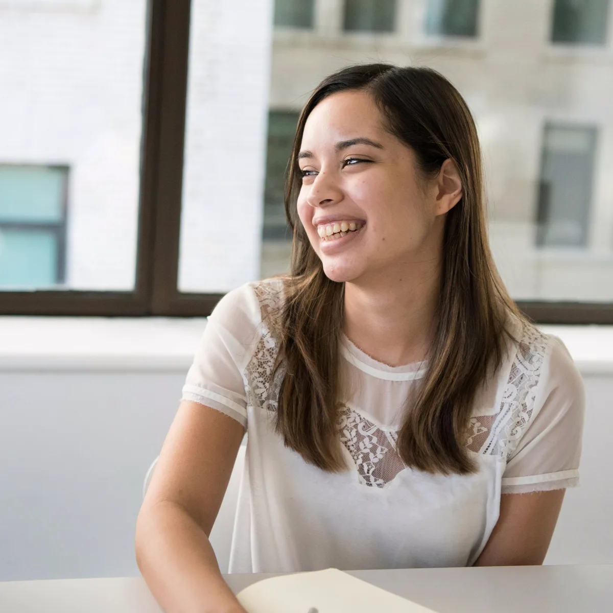 A student smiling at a desk by the window, an open notebook in front of her