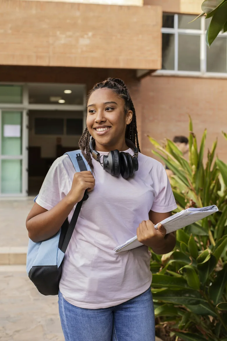 A smiling student with a backpack and notes, walking outside a campus building